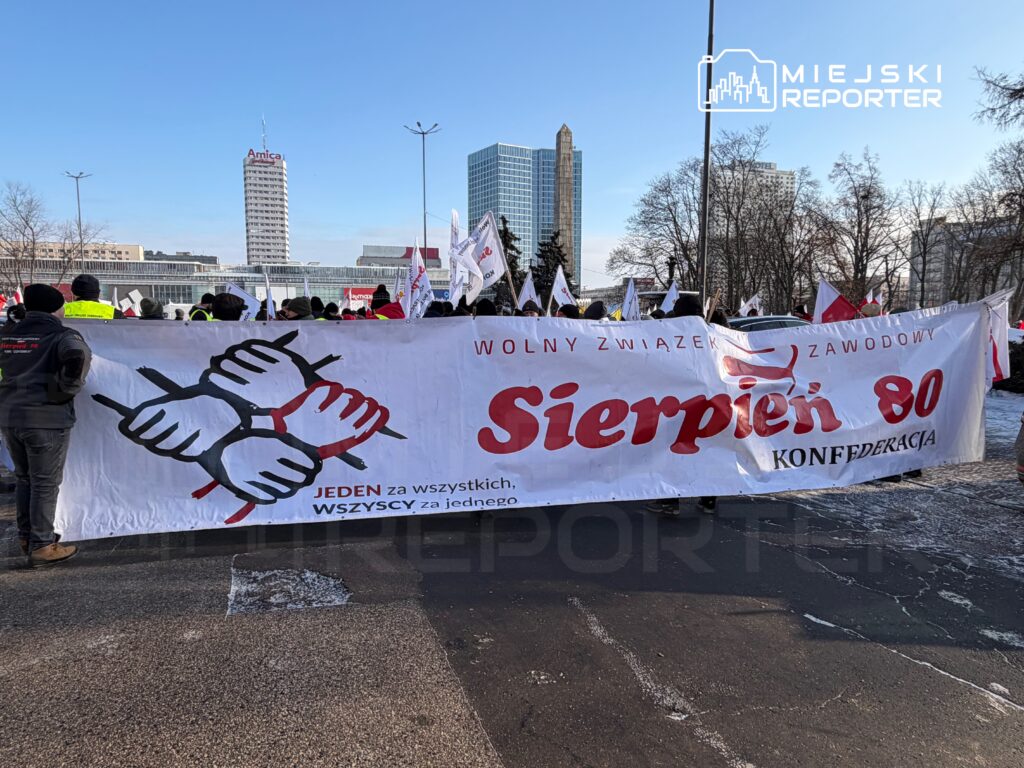 Grupa demonstrantów niosąca transparent z hasłem "Sierpień 80" oraz napisem "JEDEN za wszystkich, WSZYSCY za jednego" na tle nowoczesnych budynków.