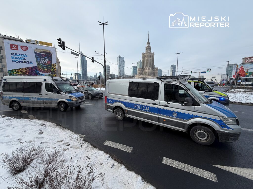 Policyjny furgon z niebieskimi oznaczeniami skręca w ruchliwym rondzie, w tle wieżowiec z zegarem.