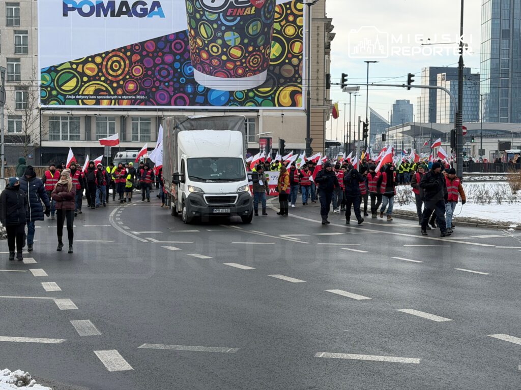Grupa protestujących z flagami Polski maszeruje ulicą, obok stoi biały samochód dostawczy. W tle nowoczesne budynki.