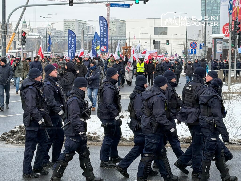 Grupa policjantów w mundurach i z hełmami patroluje ulicę w czasie demonstracji, w tle tłum z flagami i transparentami.