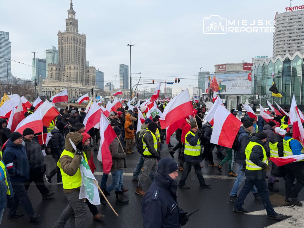 Grupa ludzi z polskimi flagami uczestniczy w demonstracji w centrum miasta, w tle widoczny wysoki budynek.