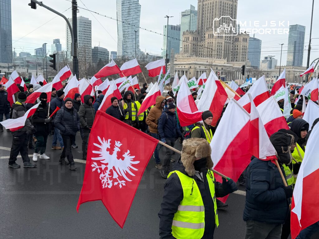 Tłum ludzi z flagami w barwach narodowych maszeruje ulicą, w tle nowoczesne wieżowce.