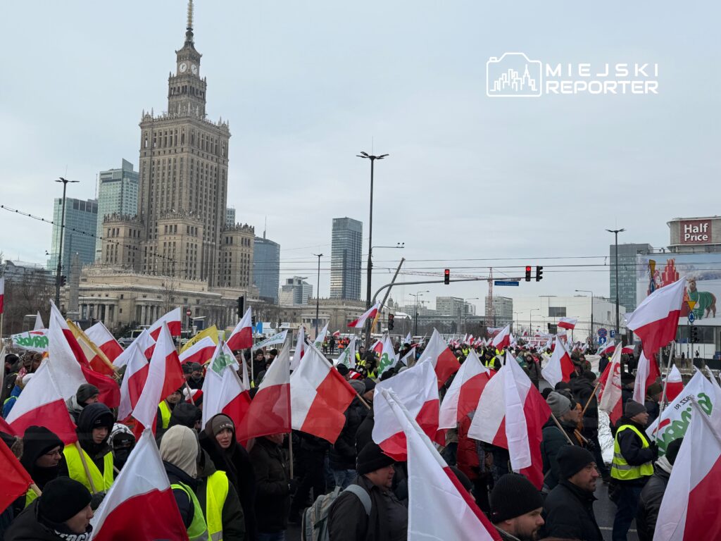 Tłum ludzi z flagami w narodowych barwach protestuje na ulicy przed wysokim budynkiem z zegarem.