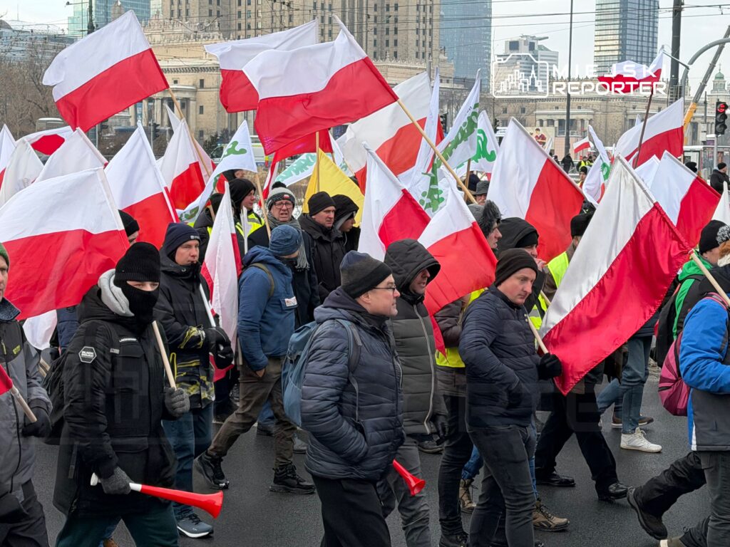 Grupa demonstrantów z flagami Polski maszeruje ulicą w mieście, w tle nowoczesne budynki.