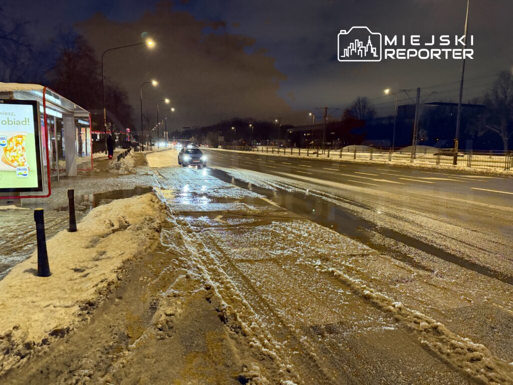 Samochód porusza się po ośnieżonej drodze w nocy, obok przystanku z reklamą na wiaty.