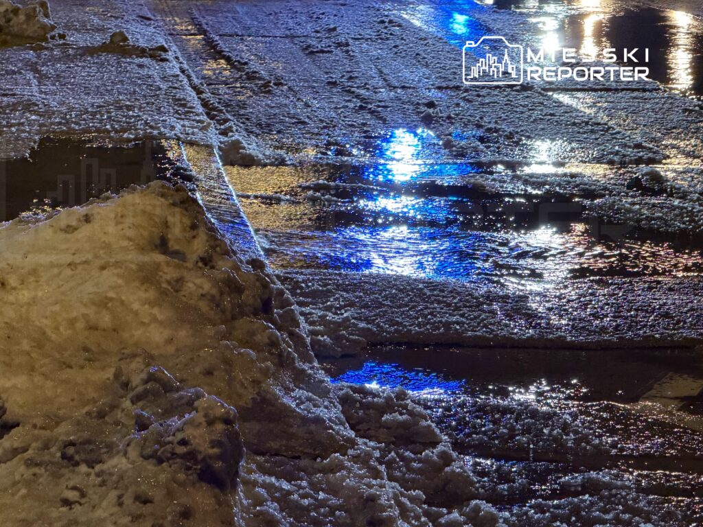 Zasypana śniegiem ulica nocą, refleksy świateł odbijają się w mokrej nawierzchni.
