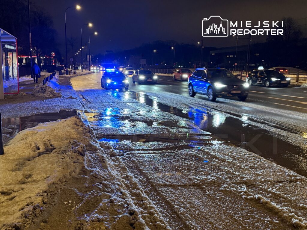 Radiowóz z niebieskimi światłami zatrzymany na oświetlonej ulicy, obok leżąca śnieg i mokra nawierzchnia.