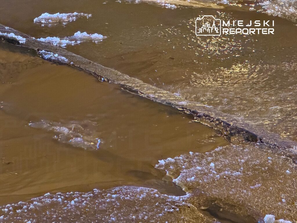 Zamrożona kałuża z lodem i wodą, w tle widoczna asfaltowa nawierzchnia.