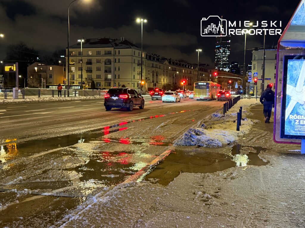 Ruchliwa ulica nocą z samochodami i śniegiem, odbicia świateł w kałużach, przechodzień idący chodnikiem.