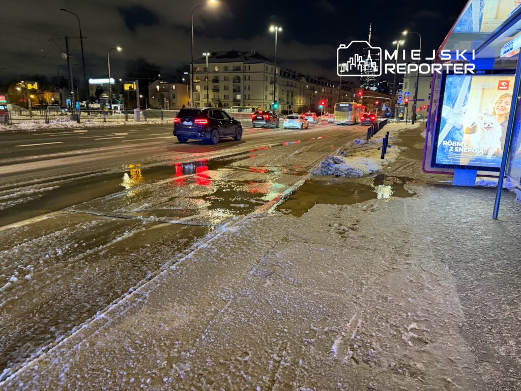 Zimowa noc na ruchliwej ulicy z odbiciem świateł w kałużach na jezdni i śniegiem na chodniku.