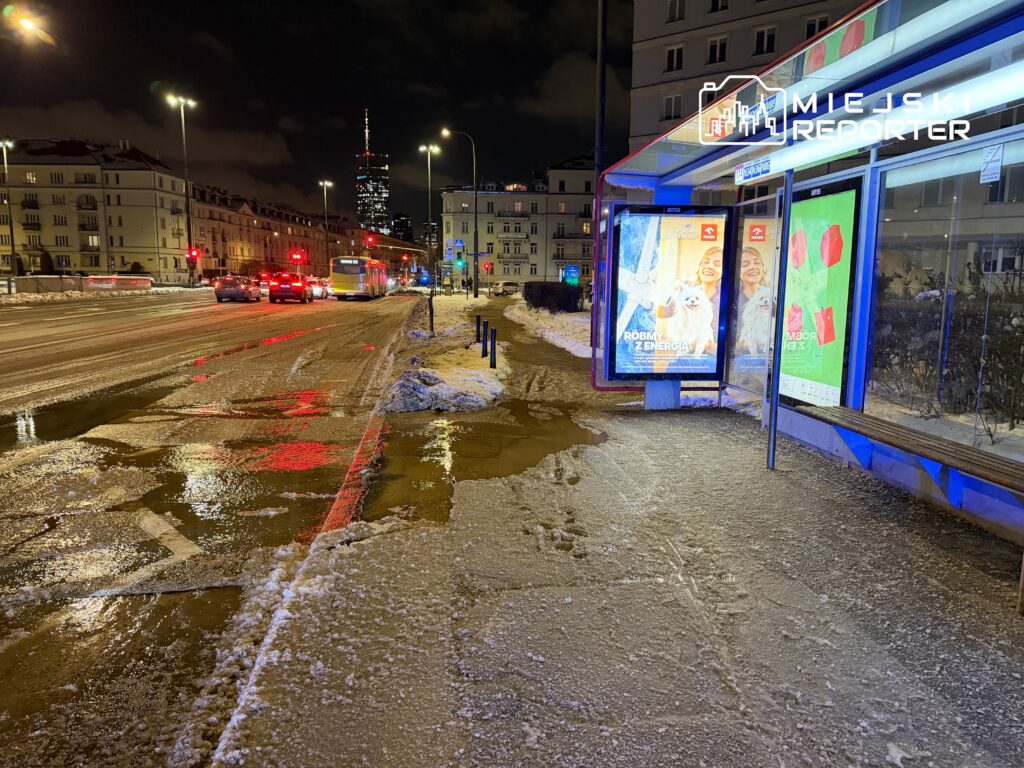 Ruchliwe skrzyżowanie uliczne nocą, z oświetlonymi budynkami, tramwajem i samochodami, na tle zaśnieżonych chodników.