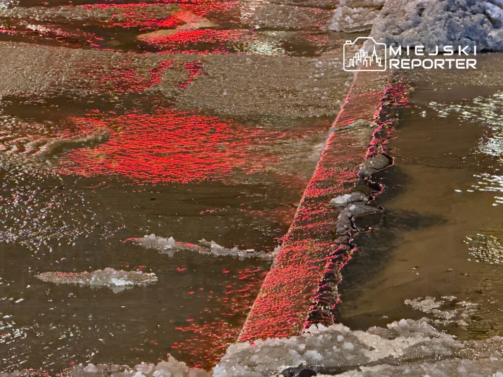 Odbicie czerwonego światła na mokrej nawierzchni ulicy z fragmentami lodu i wody.