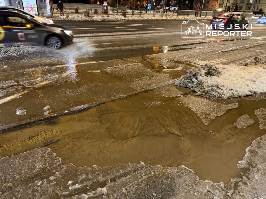 Samochód przejeżdża przez zamarzniętą ulicę, na której widać kałużę brudnej wody i resztki śniegu.