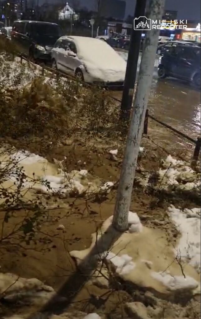 Zasypane śniegiem samochody zaparkowane przy oświetlonej ulicy, w tle widoczna woda na jezdni.