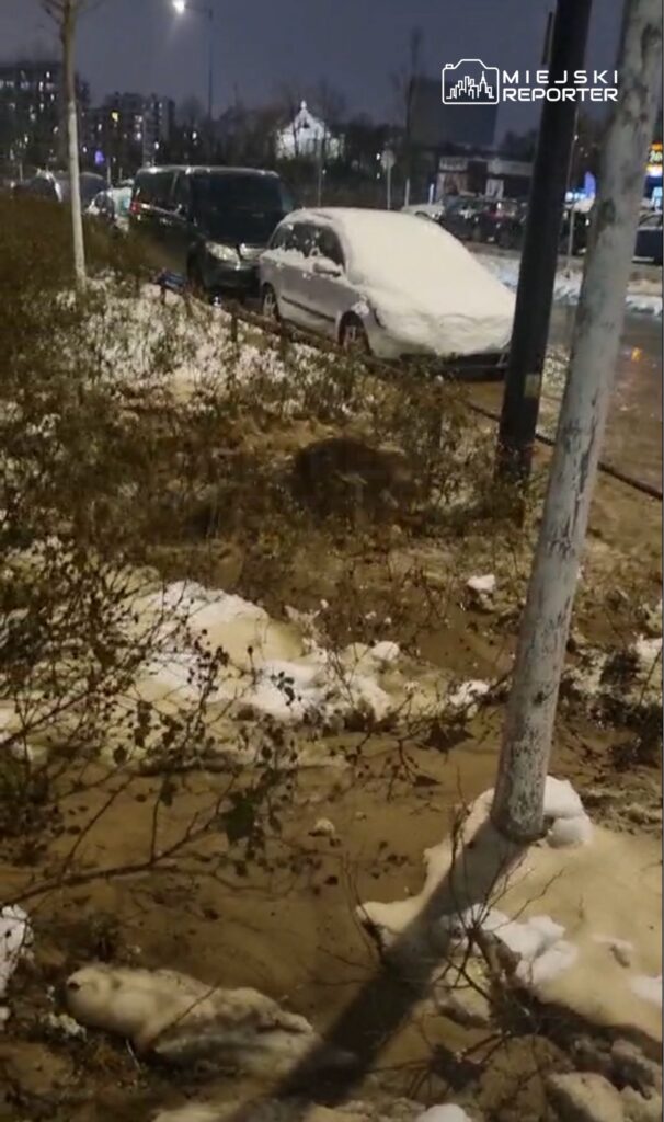 Zasypane śniegiem samochody zaparkowane wzdłuż drogi, w tle widoczne oświetlone budynki nocą.