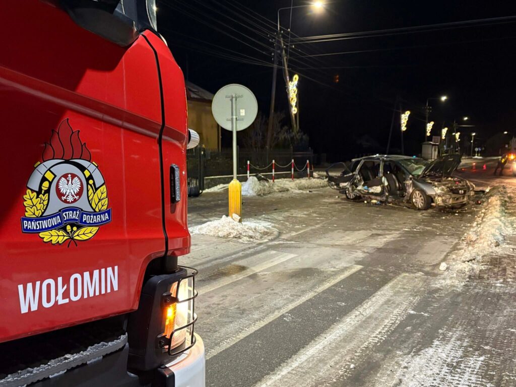 Uszkodzony samochód osobowy stoi na drodze, obok czerwony wóz strażacki z napisem „Państwowa Straż Pożarna Wołomin”.