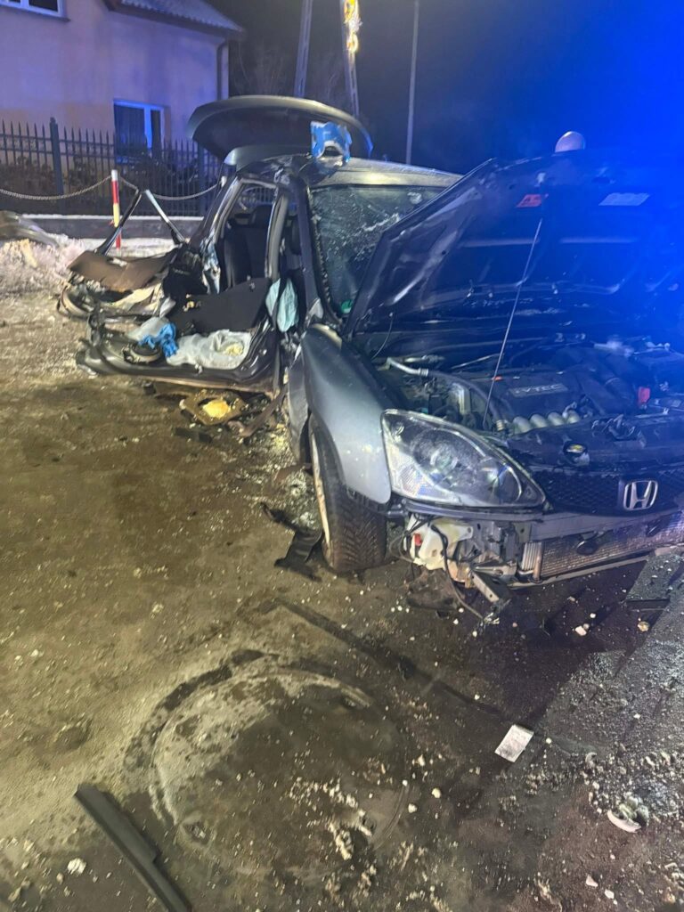 Zniszczony samochód osobowy z otwartymi drzwiami, leżący na drodze w nocy, w otoczeniu śniegu i słabych świateł.