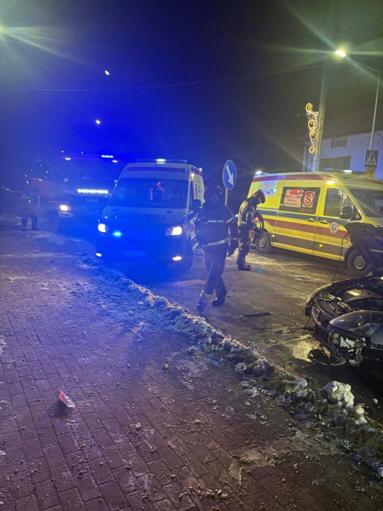 Ratownicy medyczni i strażacy pracują przy wypadku drogowym na oświetlonej ulicy, obok uszkodzonego samochodu.