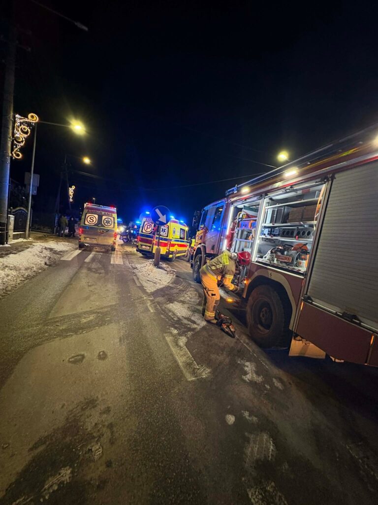 Strażak w żółtym mundurze sprawdza sprzęt przy wozie strażackim na oświetlonej, zaśnieżonej ulicy. W tle widoczne karetki.