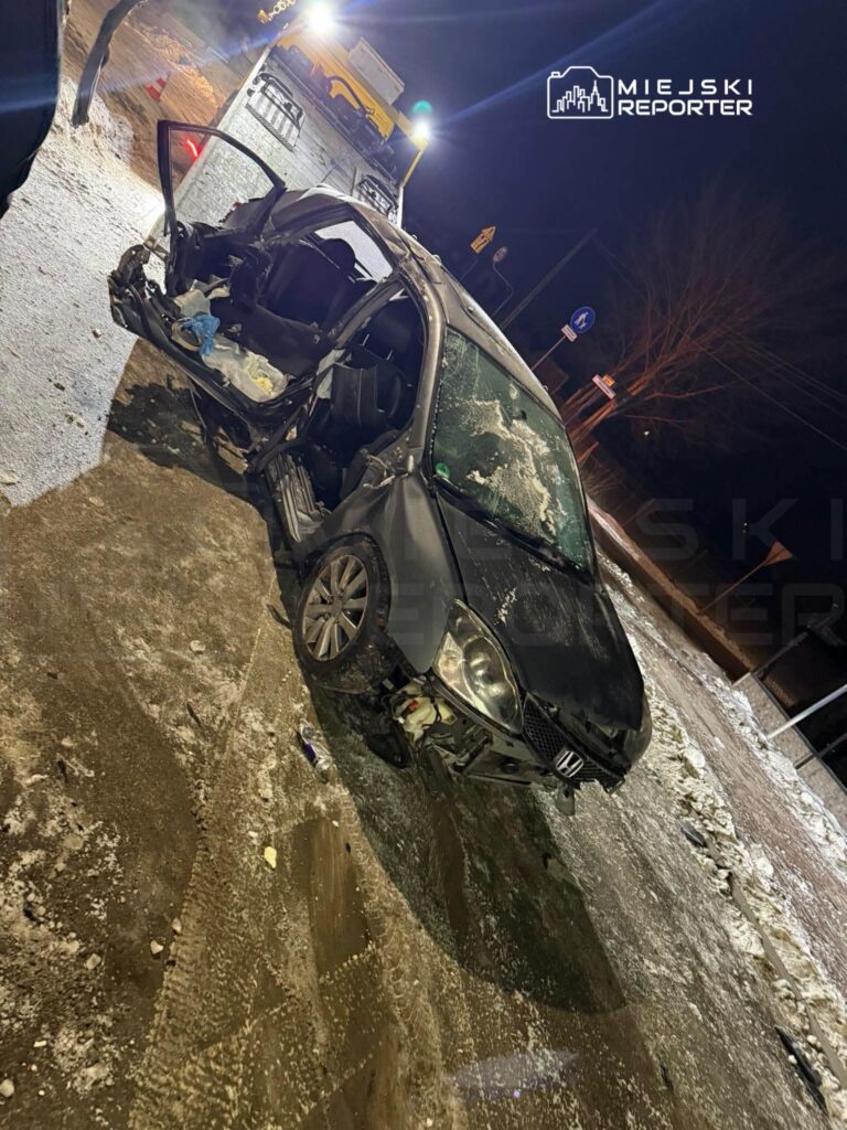 Zniszczony samochód osobowy stoi na ośnieżonej drodze, obok niego widoczny jest laweta oraz znaki drogowe.