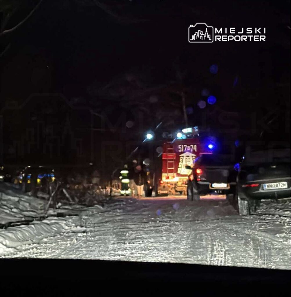 Strażacy pracują przy wozie strażackim na ośnieżonej drodze w nocy, obok zaparkowanych samochodów.