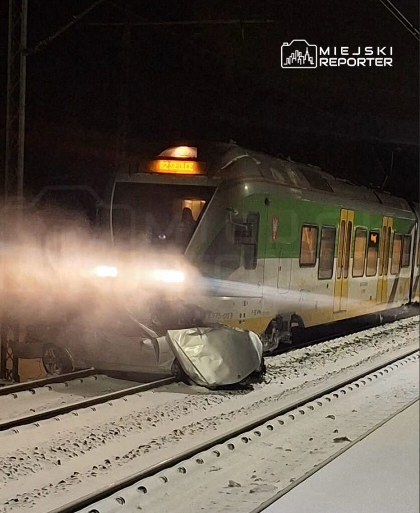 Pociąg towarowy zatrzymany na torach, obok uszkodzone auto z wgniecionym przodem, otoczone śniegiem.