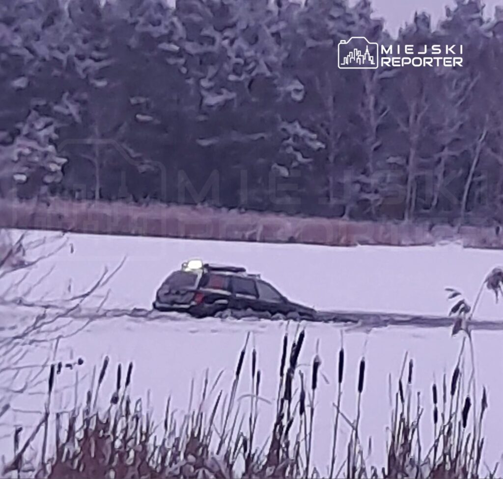 Samochód z włączonymi światłami, częściowo zanurzony w śniegu na zamarzniętym jeziorze, otoczony zimowym krajobrazem.
