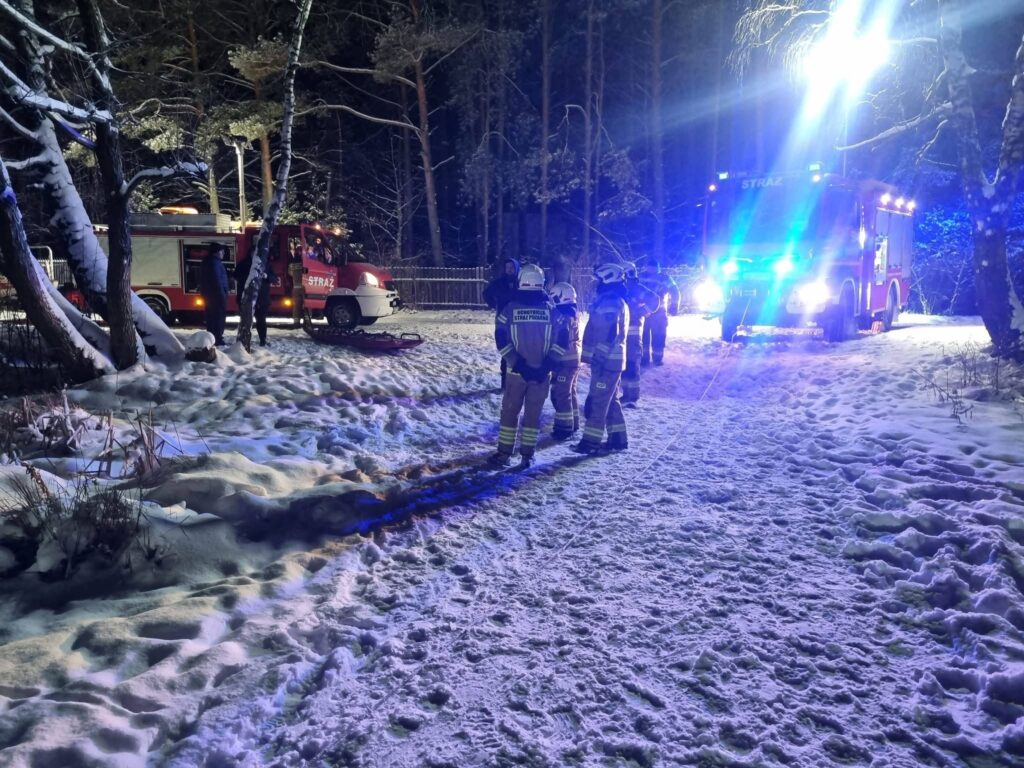 Grupa strażaków w mundurach omawia sytuację obok wozu strażackiego na ośnieżonej leśnej drodze nocą.