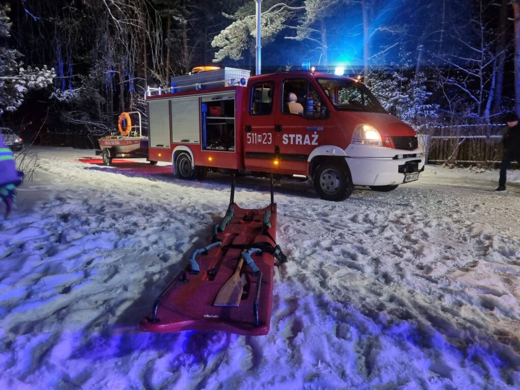Strażacy przy wozie strażackim na ośnieżonej drodze, przygotowujący nosze do akcji ratunkowej w lesie.
