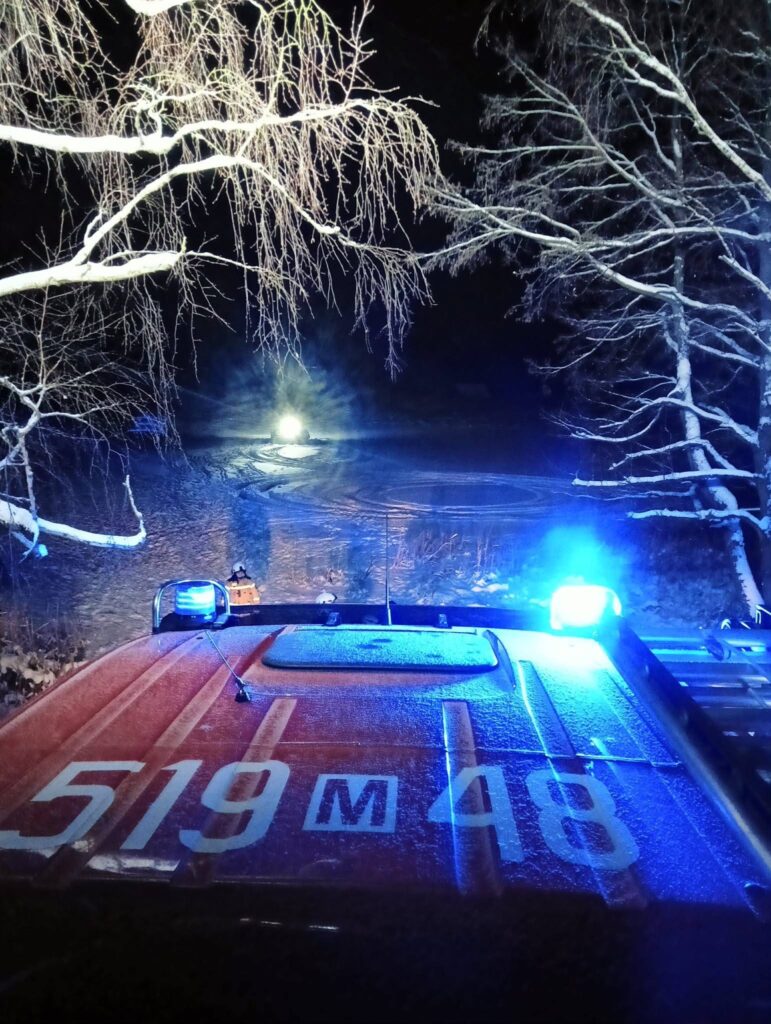 Widok na ośnieżoną drogę nocą, z niebieskimi światłami pojazdu ratunkowego na pierwszym planie.