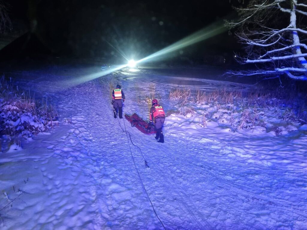 Dwóch ratowników w odblaskowych kamizelkach ciągnie nosze po zaśnieżonym terenie w nocy, oświetlonym przez reflektory.