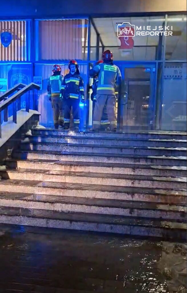 Grupa strażaków w mundurach i hełmach stoi na schodach przed budynkiem, oświetlonym niebieskimi światłami.