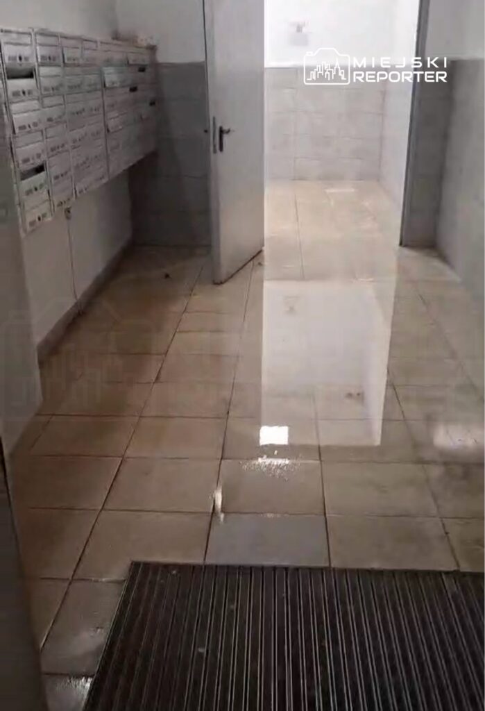 Podłoga w pomieszczeniu z pocztowymi skrzynkami jest zalana wodą, a ściany są wykończone w jasnych kolorach.