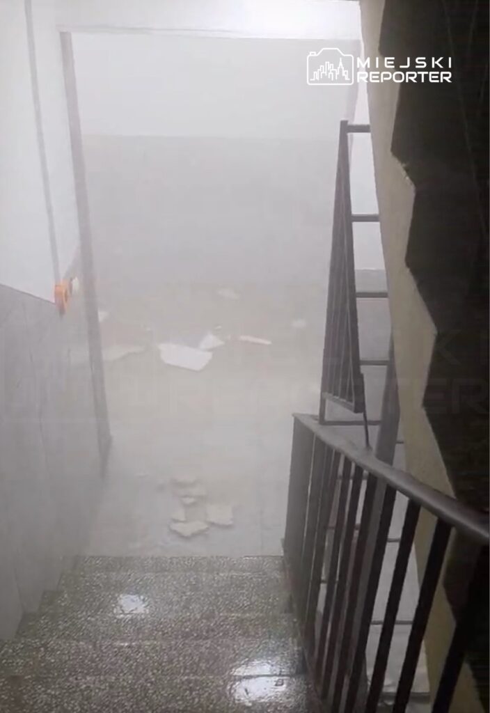 Zamglony korytarz z mokrą podłogą i porozrzucanymi papierami, widoczna metalowa balustrada schodów.