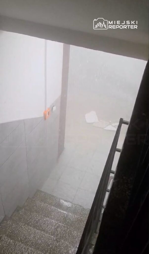 Intensywna ulewa zalewa schody w budynku, widoczność ograniczona przez deszcz i mgłę.