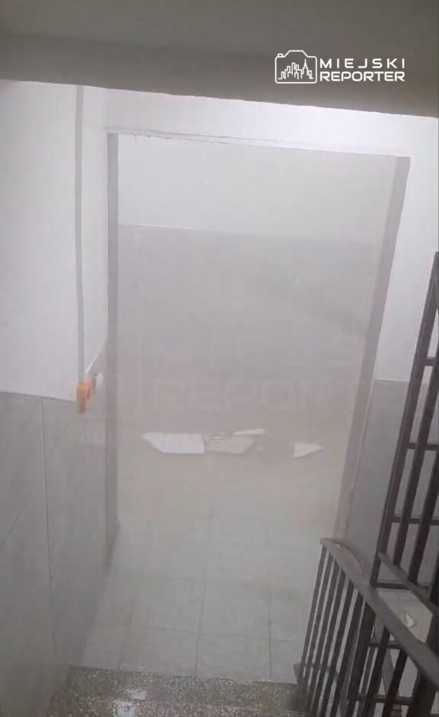 Zalany korytarz w budynku, z widocznymi zniszczeniami na podłodze i wilgocią na ścianach.