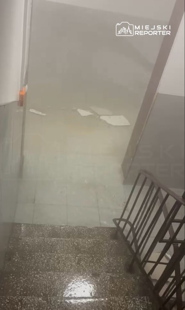 Woda zalewająca schody w budynku, z widocznymi plamami na podłodze oraz fragmentami materiałów budowlanych.