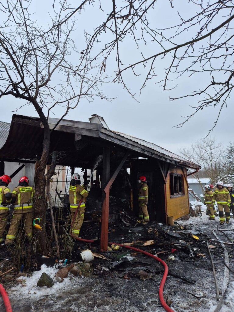 Grupa strażaków w specjalistycznych strojach gasi pożar zniszczonego budynku w zimowym otoczeniu.