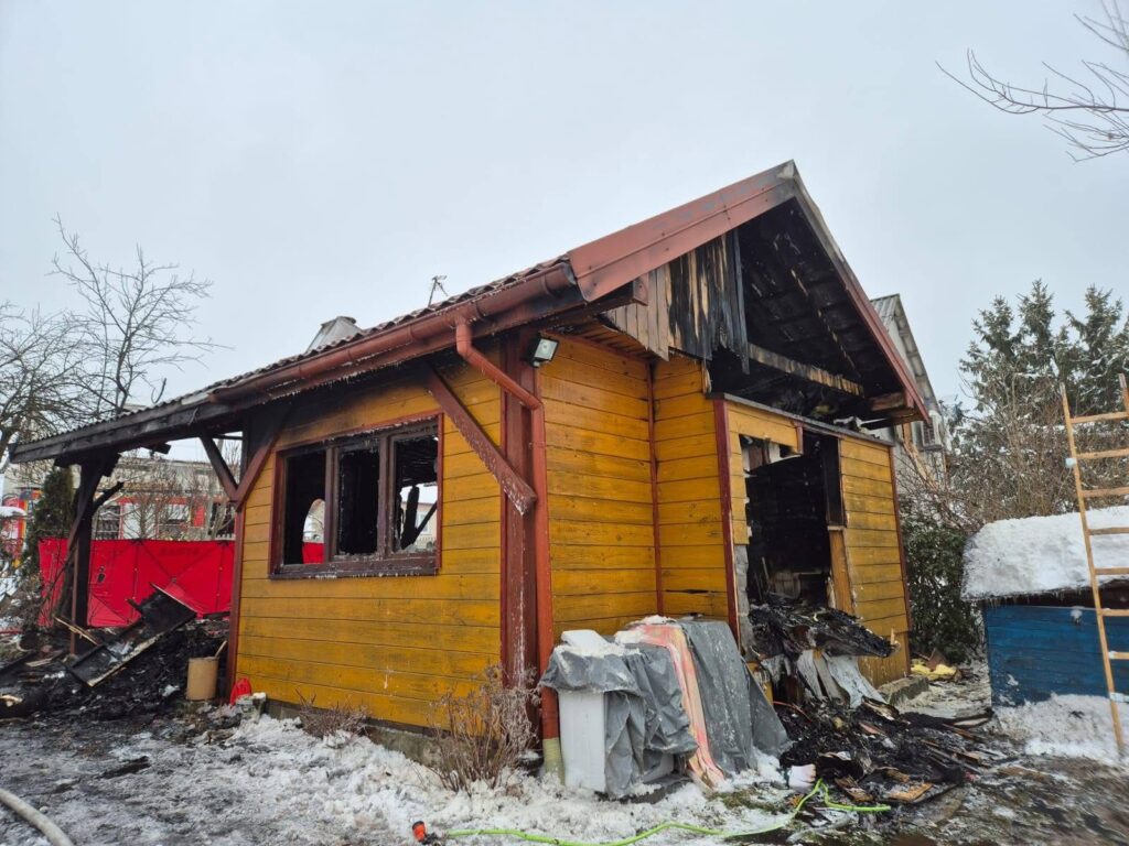 Spalony drewniany domek z uszkodzonymi oknami, w otoczeniu śniegu i zniszczeń po pożarze.