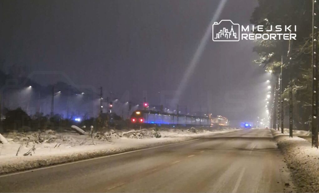 Pociąg towarowy przejeżdża w nocy po zaśnieżonej trasie, otoczony przez oświetlone latarnie.