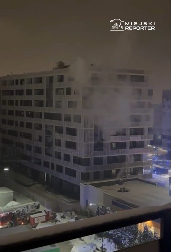 Dym wydobywający się z okna nowoczesnego budynku mieszkalnego w nocy, w otoczeniu zaśnieżonej ulicy i pojazdów ratunkowych.