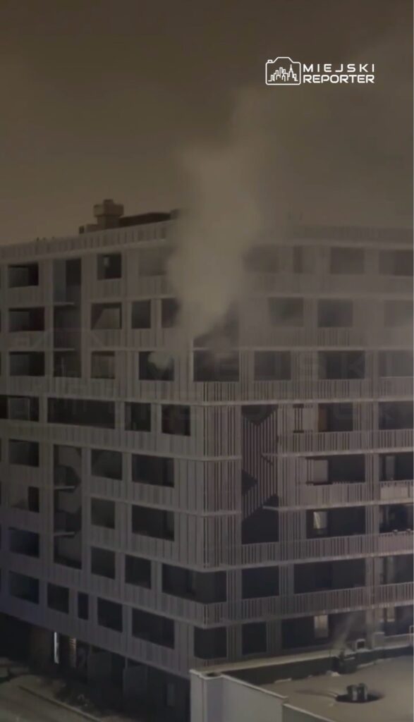 Z budynku wydobywa się dym, a na elewacji widoczne są otwarte okna w nowoczesnej architekturze.