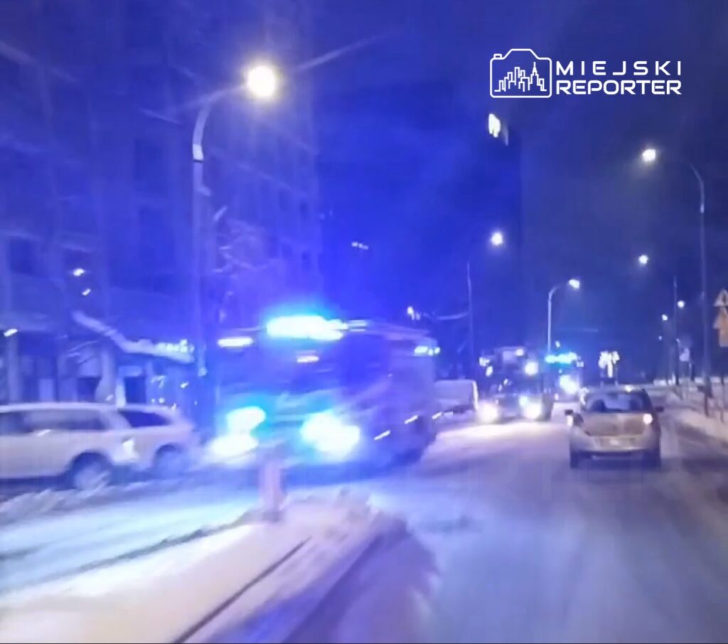 W nocy na oświetlonej ulicy przejeżdżają pojazdy ratunkowe z włączonymi syrenami i niebieskimi światłami.