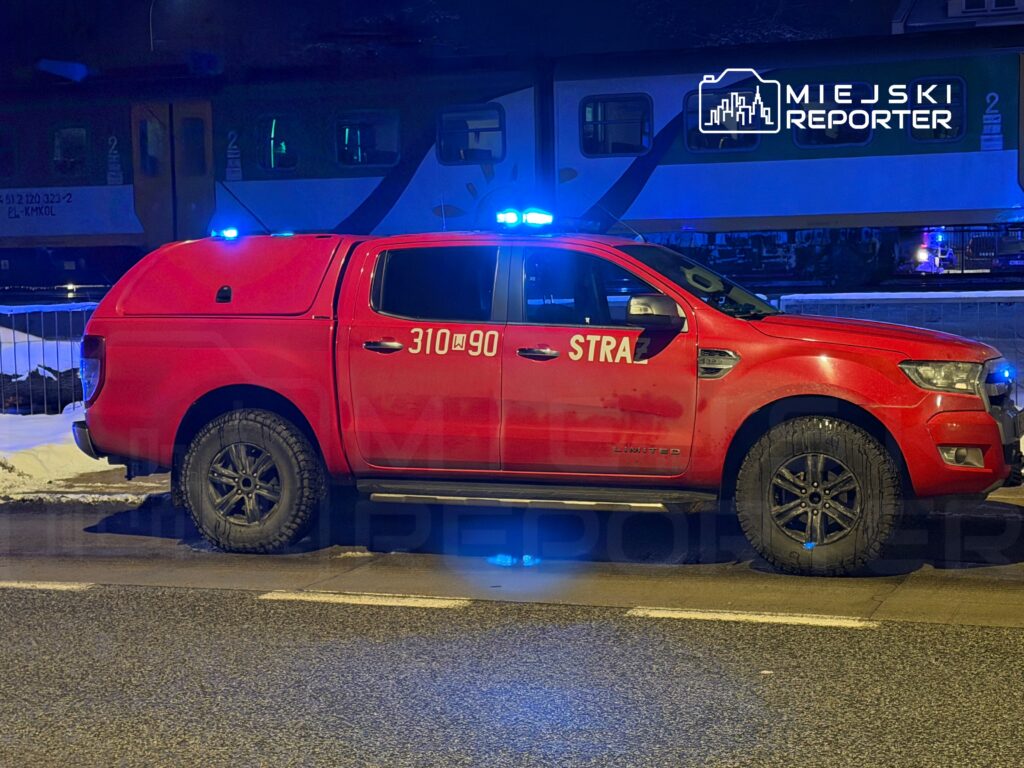 Czerwony samochód straży pożarnej z niebieskimi światłami stojący na ulicy obok torów kolejowych w nocy.