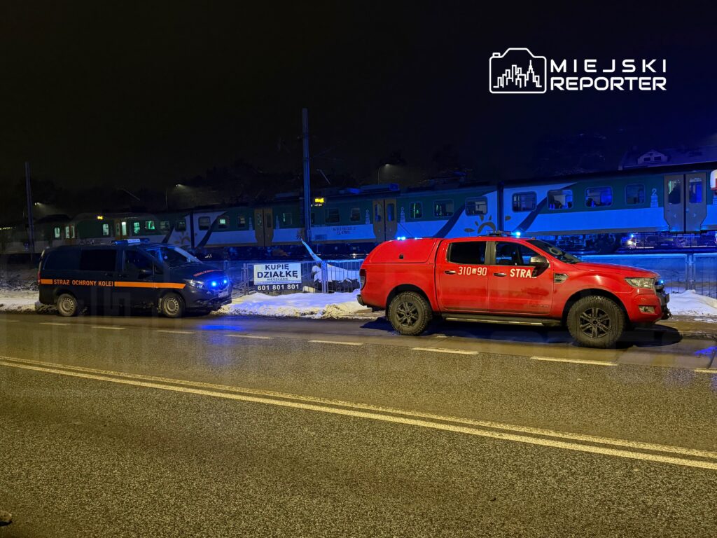 W nocy, dwa pojazdy służbowe stoją obok torów kolejowych; czarny van Straży Ochrony Kolei i czerwony samochód strażacki.