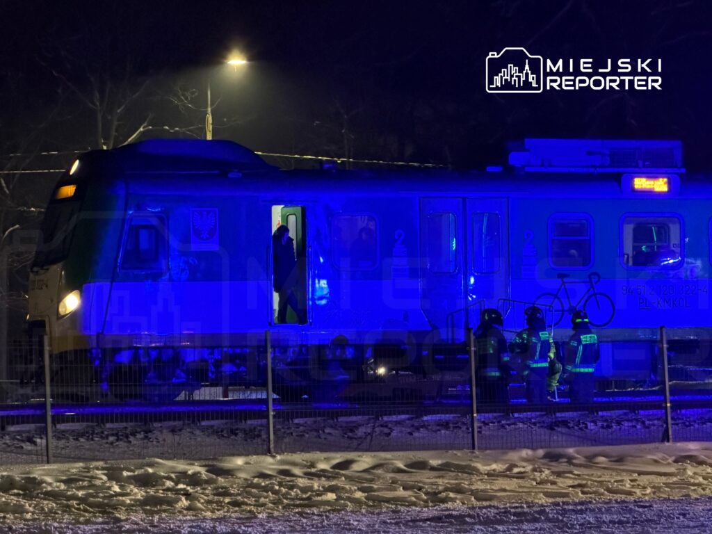 Pociąg zatrzymany na stacji, przy otwartych drzwiach, obok grupy strażaków w mundurach, otoczenie oświetlone niebieskim światłem.