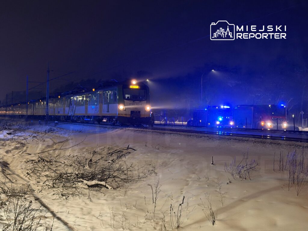 Pociąg towarowy zatrzymany przy torach w zimowym otoczeniu, obok niebieskie światła wozów strażackich.