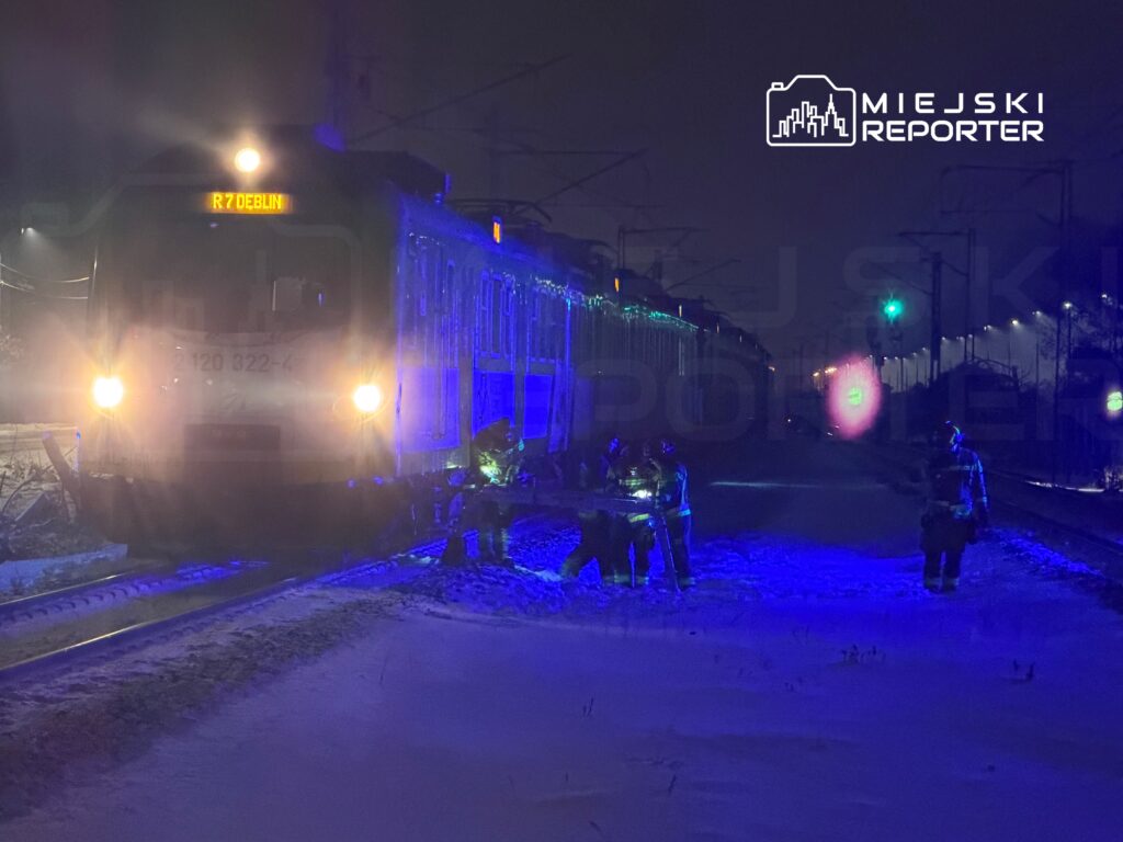 Pociąg z żółtym wyświetlaczem "R7 DEBLIN" zatrzymany na zaśnieżonej stacji, obok pracujący zespół ratunkowy w niebieskich mundurach.