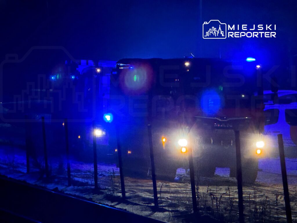 W nocy, w oświetlonych niebieskim światłem pojazdach służb ratunkowych, stojących za ogrodzeniem.