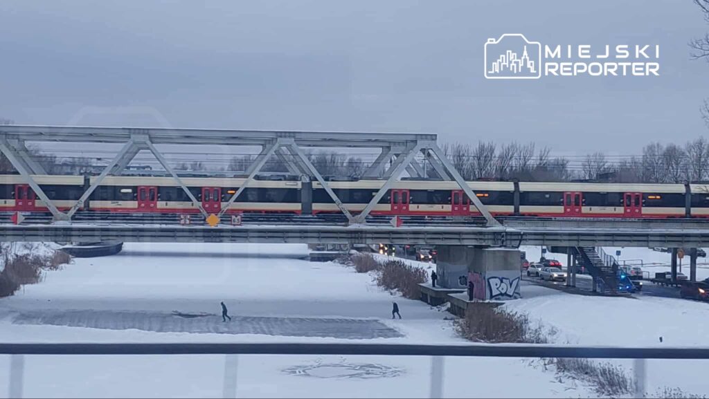 Pociąg przejeżdża przez stalowy most nad zamarzniętą rzeką, obok dwóch osób spacerujących po śniegu.